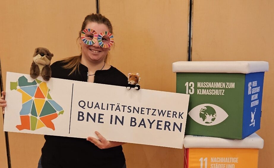 Frau mit bunter Brille h&auml;lt Schild "Qualit&auml;tsnetzwerk BNE in Bayern", neben W&uuml;rfeln mit Nachhaltigkeitszielen und Stofftier, auf Holzfu&szlig;boden vor hellem Hintergrund.