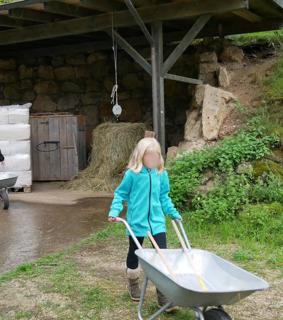 Ein Kind mit blonden Haaren schiebt eine leere Schubkarre drau&szlig;en auf einem Bauernhof, im Hintergrund Heuballen und ein Holzschuppen.