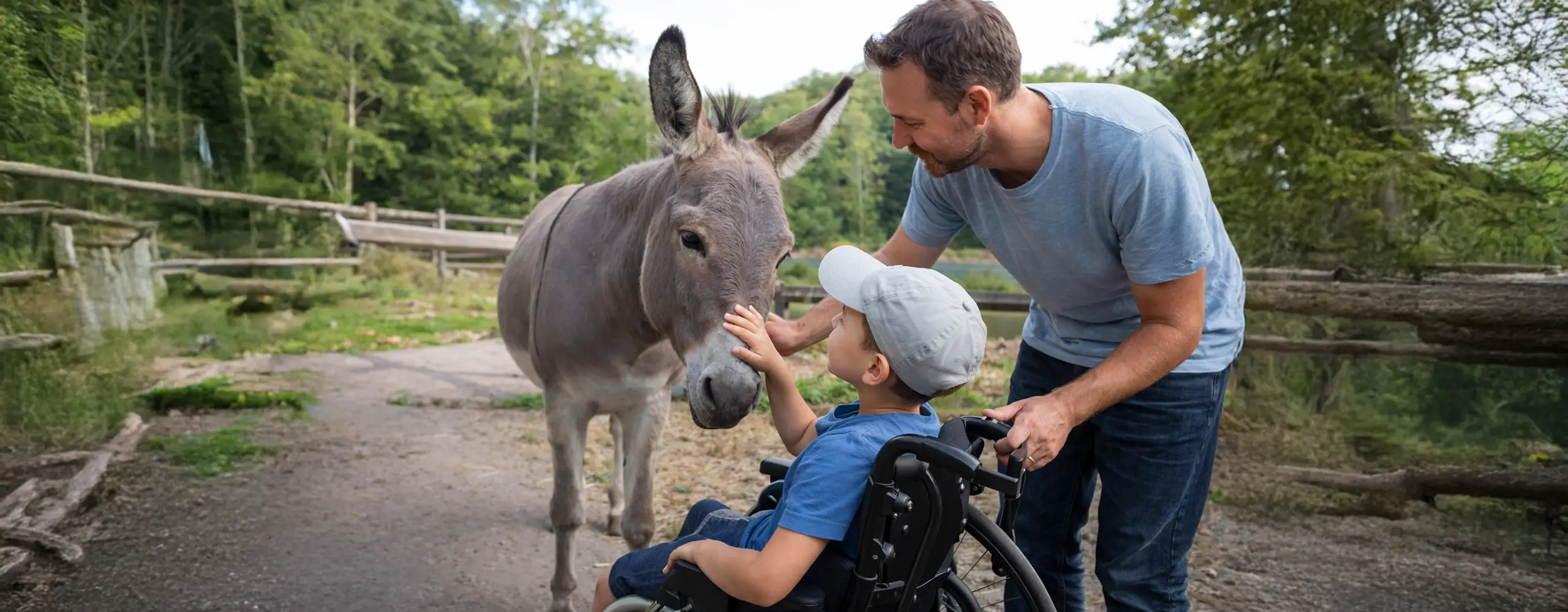 Ein Junge im Rollstuhl streichelt einen Esel, während ein Mann ihn lächelnd unterstützt. Sie sind draußen in einer natürlichen Umgebung.