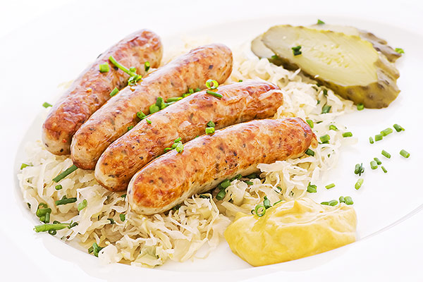 Vier Bratwürste auf Sauerkraut mit Schnittlauch bestreut, dazu Senf und Gewürzgurken auf einem weißen Teller.