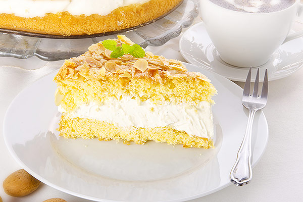 Ein Stück Bienenstichkuchen mit cremiger Füllung und Mandeln auf einem weißen Teller, daneben eine Tasse und ein Kuchen im Hintergrund.