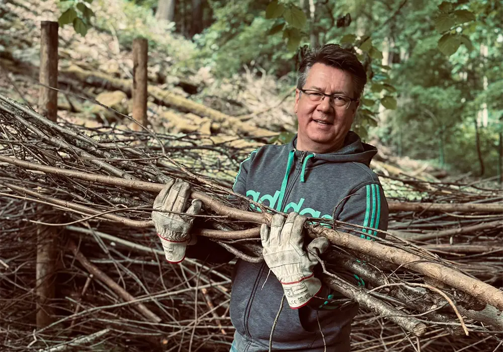 Eine Person mit Handschuhen trägt große Zweige im Freien, umgeben von Bäumen und Ästen.