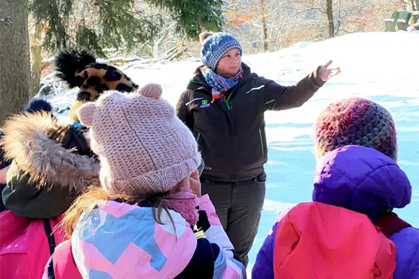Eine Person im Wintermantel erklärt einer Gruppe von Kindern im Freien, umgeben von Schnee. Die Kinder tragen bunte Winterkleidung und Mützen.