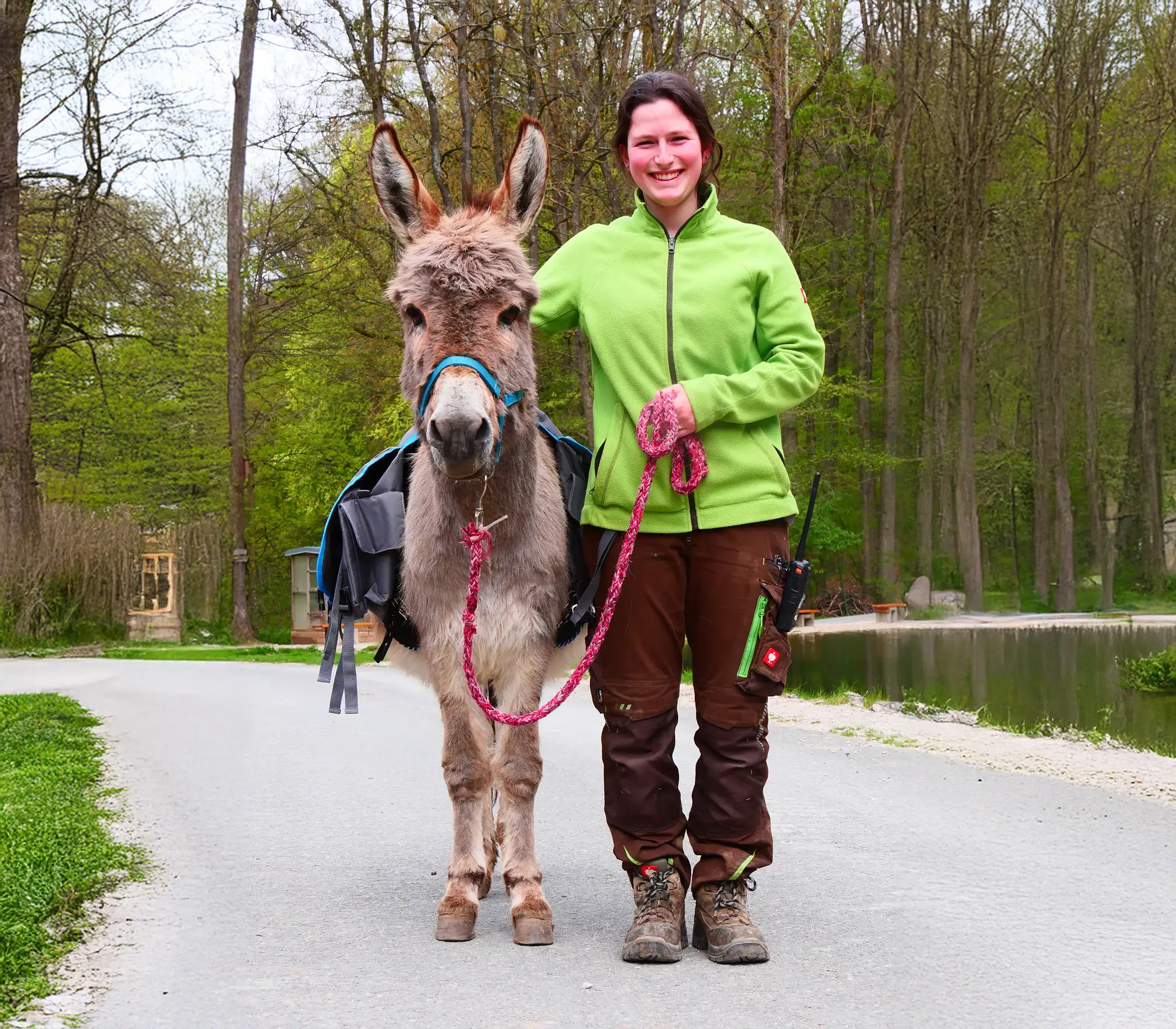Eine Frau in gr&uuml;ner Jacke steht im Freien auf einem Weg neben einem Esel an der Leine, Wald im Hintergrund.