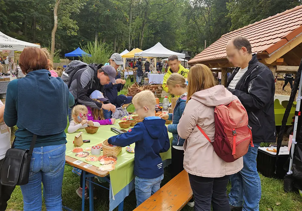 Menschen, darunter Kinder und Erwachsene, stehen an einem Tisch im Freien bei einer Veranstaltung mit Zelten im Hintergrund und besch&auml;ftigen sich mit Bastelarbeiten.