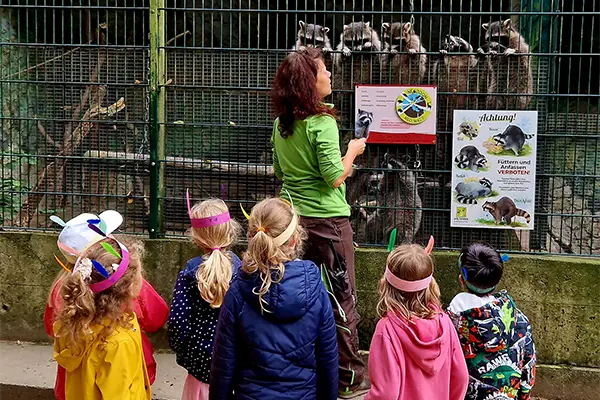 Eine Frau erklärt einer Gruppe von Kindern vor einem Zaun mit Tieren und Infotafeln etwas. Die Kinder tragen bunte Jacken und Tiermasken.
