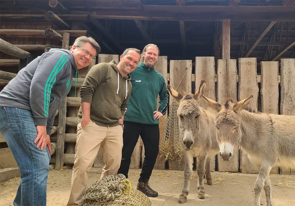 Drei Männer posieren neben zwei Eseln vor einem Holzzaun in einem ländlichen Umfeld und lächeln in die Kamera.