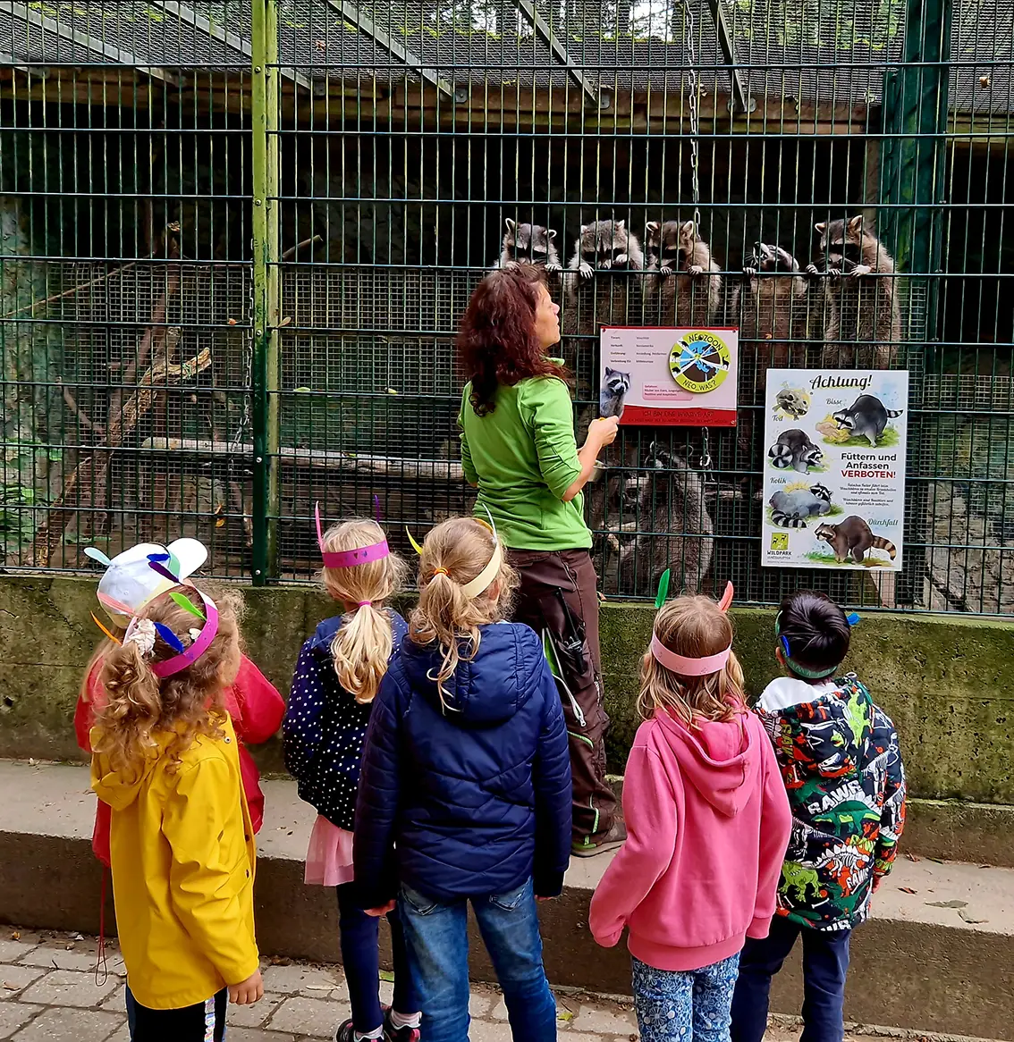 Eine Frau zeigt einer Gruppe von Kindern Tiere in einem Zoo, w&auml;hrend sie vor einem Gehege mit Informationstafeln stehen.