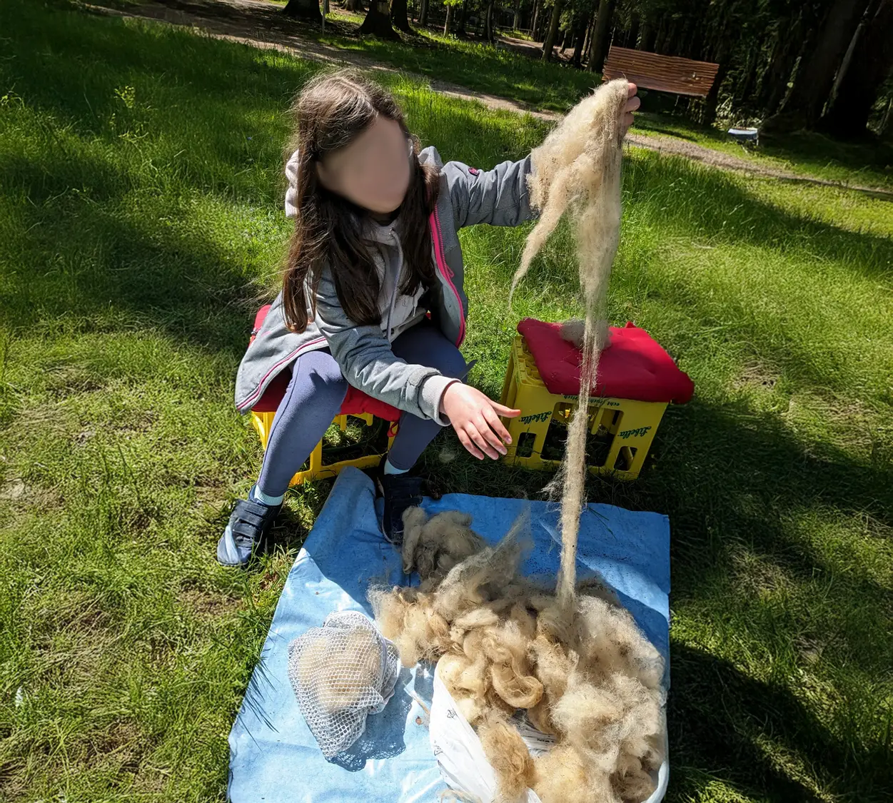 Ein M&auml;dchen sitzt drau&szlig;en auf einer Decke und verarbeitet Wolle, umgeben von Natur und gr&uuml;ner Wiese.