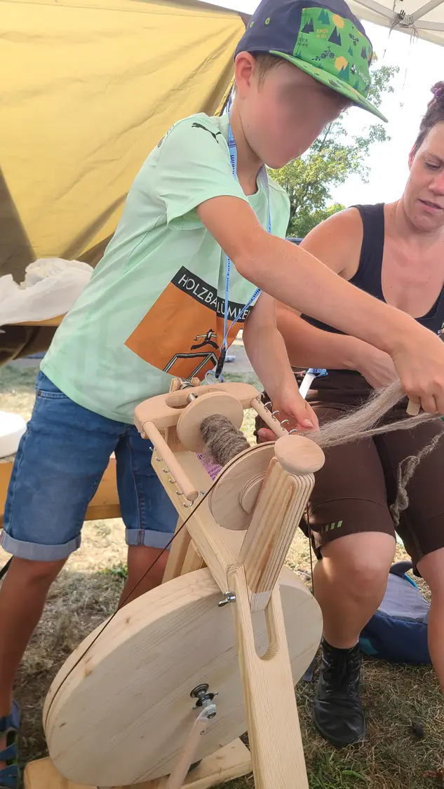 Zwei Personen bedienen ein Spinnrad und verarbeiten Fasern zu Garn, eine Person zeigt und hilft dabei.