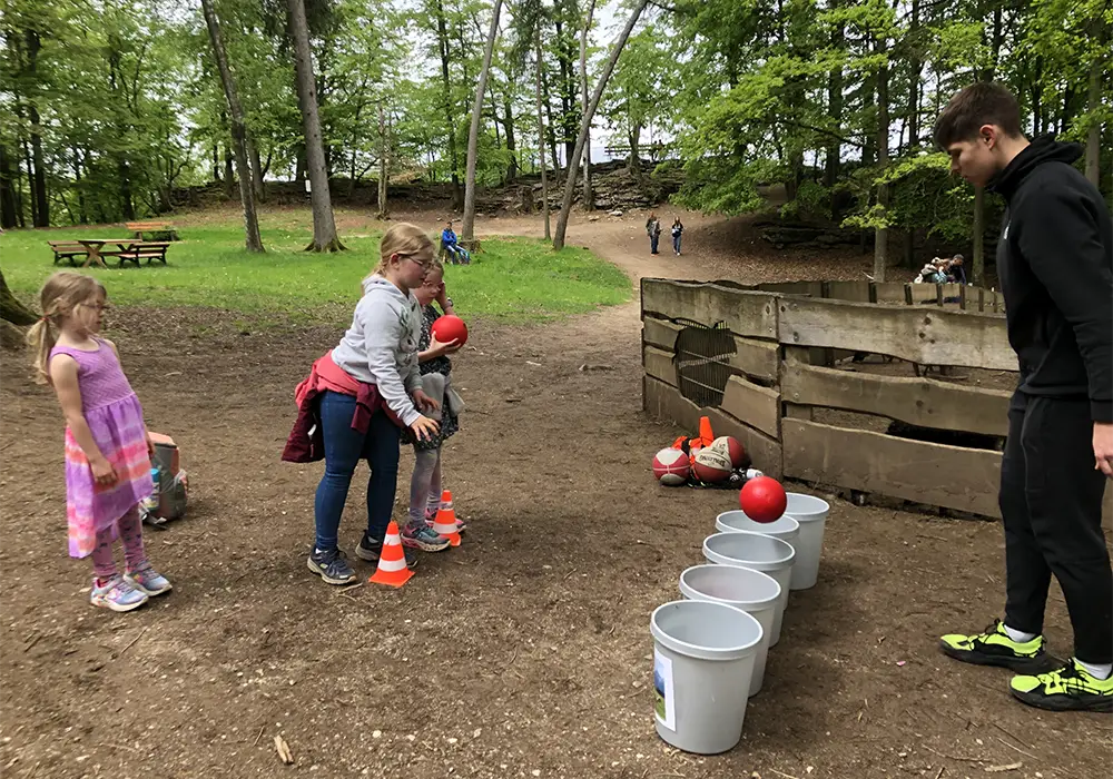 Eine Frau hilft einem Kind beim Werfen eines Balls in einen Eimer im Freien, umgeben von B&auml;umen und mehreren Eimern auf dem Boden.