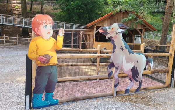 Pappfigur eines Kindes und einer Ziege vor einem Holzzaun auf einem Bauernhof, im Hintergrund stehen Stallungen und Bäume.