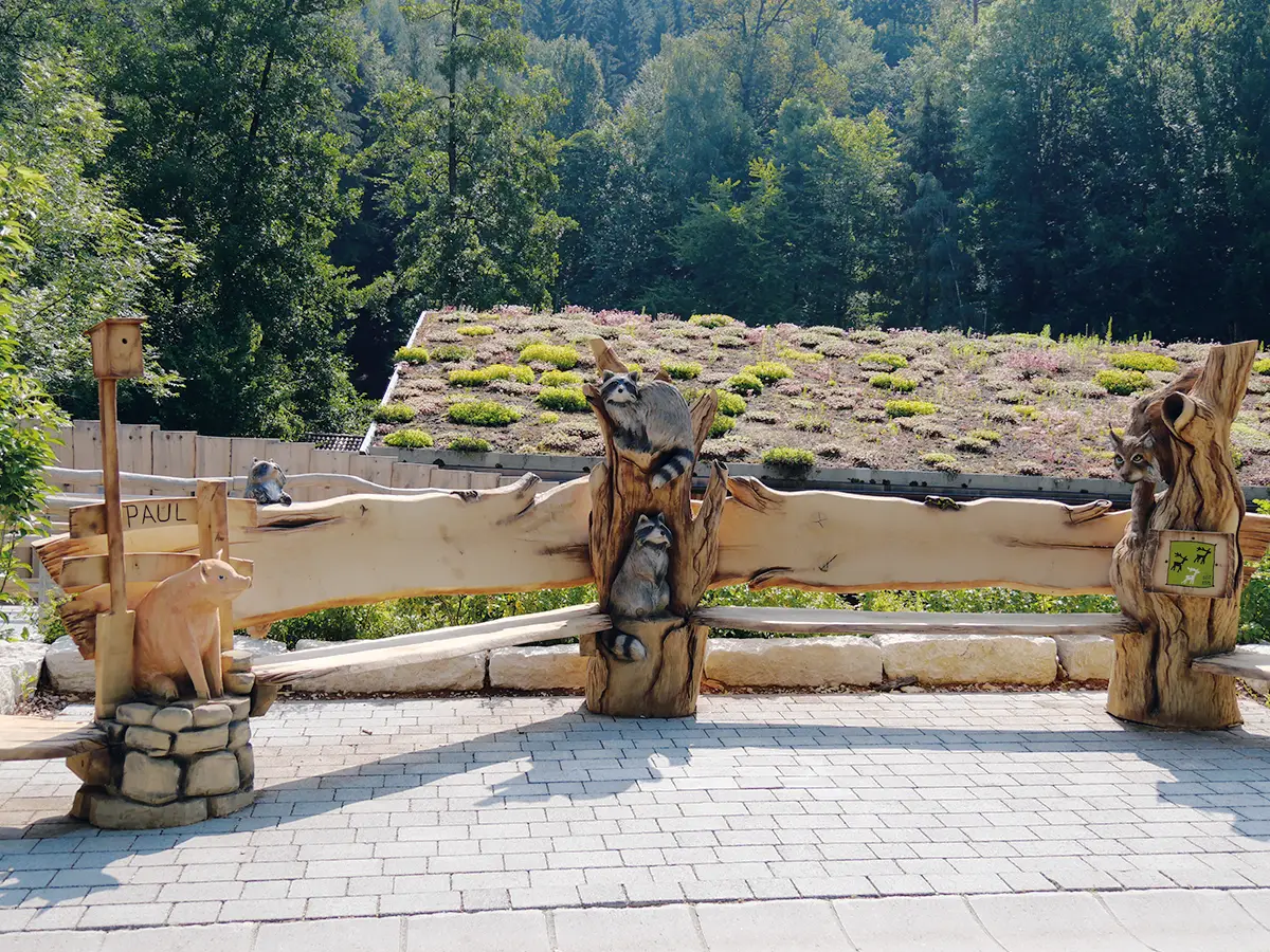 Holzzaun aus massiven, naturbelassenen Baumstämmen vor einem mit Moos bedeckten Dach, im Hintergrund bewaldete Hügel und grüne Bäume.
