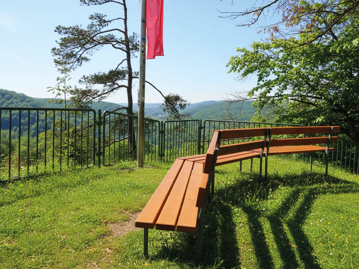 Gebogene Holzbänke auf einer Wiese mit Blick auf bewaldete Hügel, ein Zaun und eine rote Fahne im Hintergrund bei sonnigem Wetter.