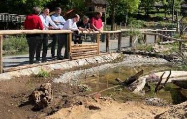 Eine Gruppe von Menschen steht an einem Holzzaun und blickt auf ein kleines, naturnahes Wasserbecken in einer grünen Außenanlage.