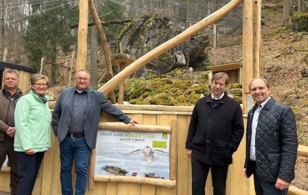 Drei Männer stehen vor einem Holzgehege mit Infotafel im Freien, dahinter Felsen und Natur.