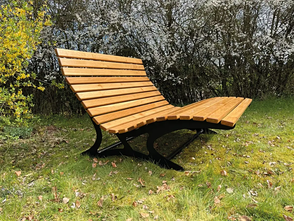 Moderne Holzliegebank mit Metallgestell steht auf einer Wiese vor dichten Büschen.