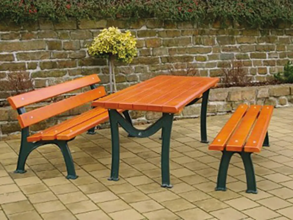 Holztisch mit zwei Holzbänken steht auf einem gepflasterten Hof vor einer Steinmauer, im Hintergrund ist eine Topfpflanze zu sehen.