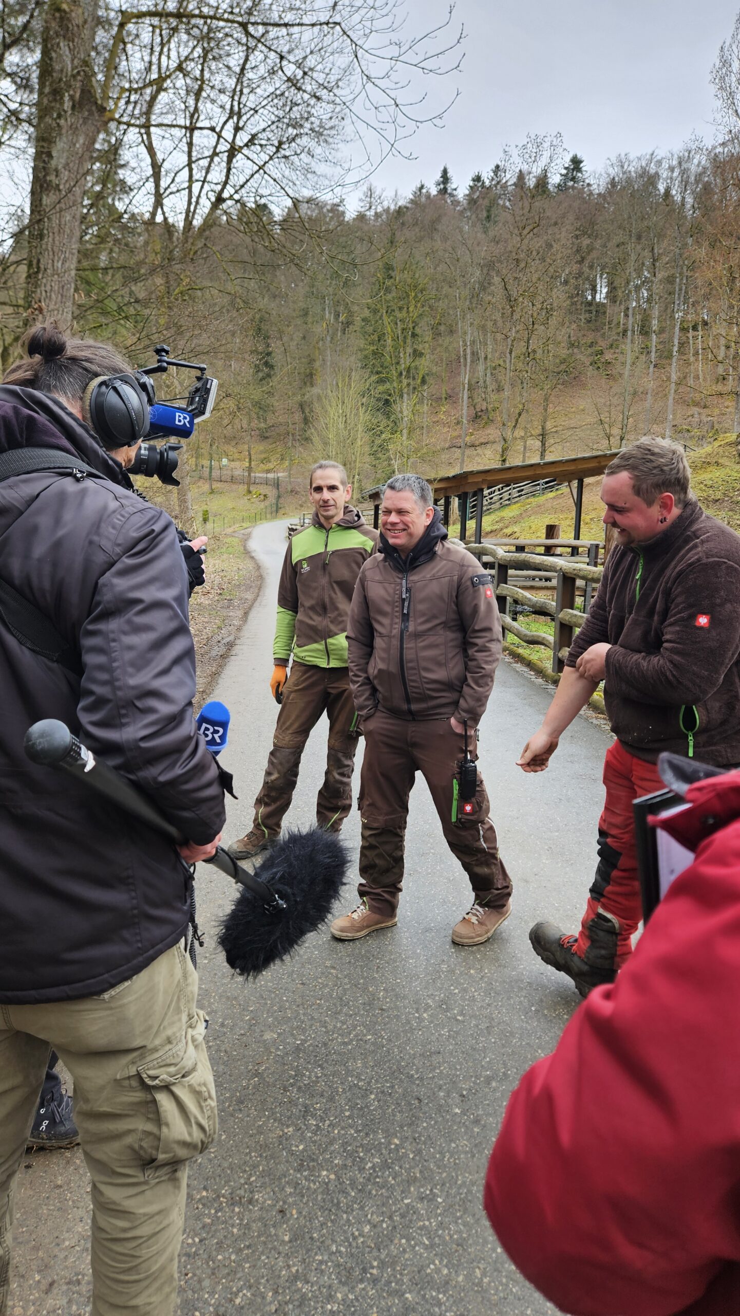 Kamerateam filmt drei M&auml;nner in Outdoor-Kleidung auf einem Waldweg, umgeben von B&auml;umen im Hintergrund. Die Stimmung wirkt locker und freundlich.
