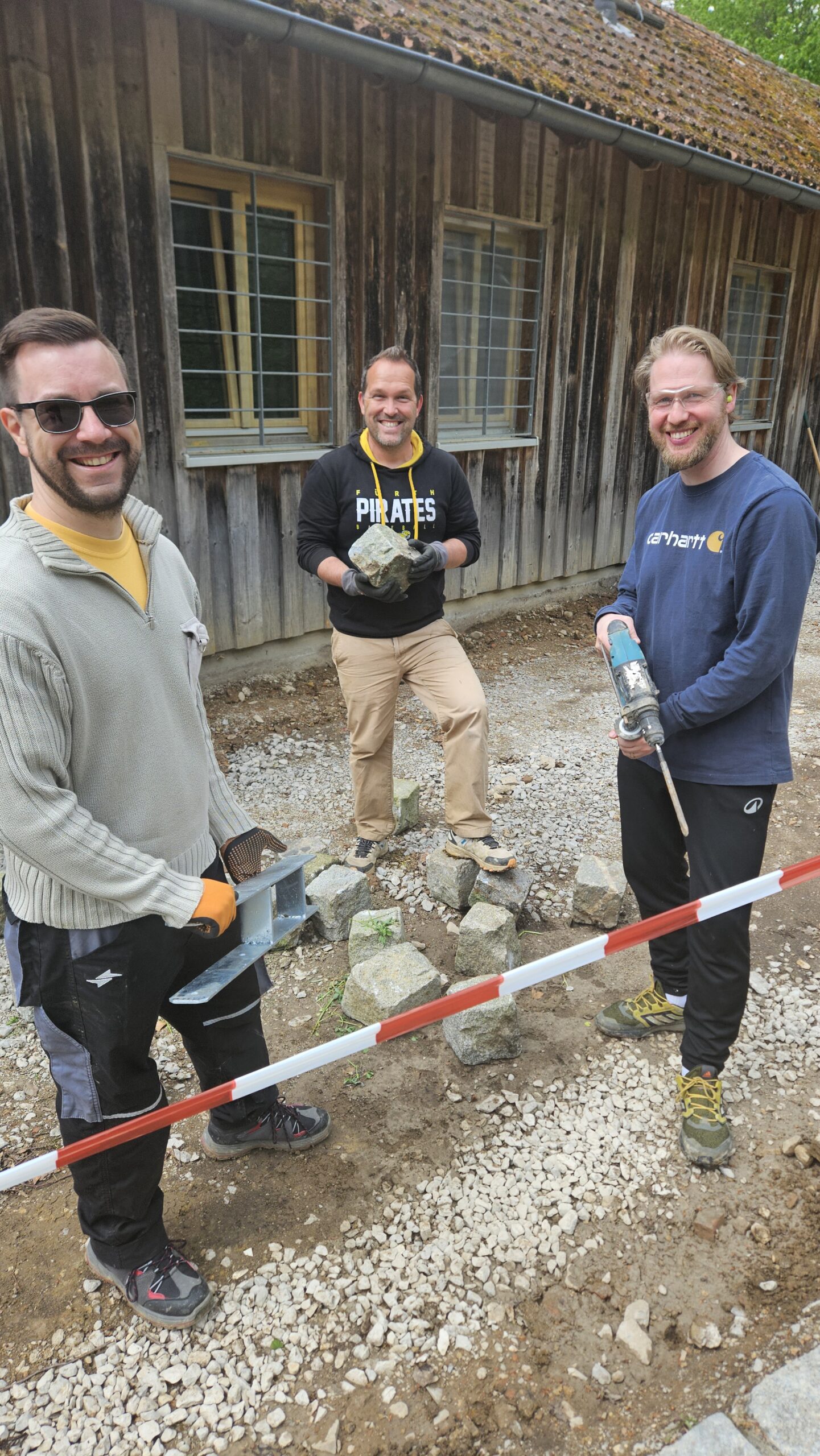 Drei Männer arbeiten draußen mit Werkzeugen und Steinen vor einem alten Holzhaus, sie lächeln in die Kamera und tragen Arbeitskleidung und Schutzbrillen.