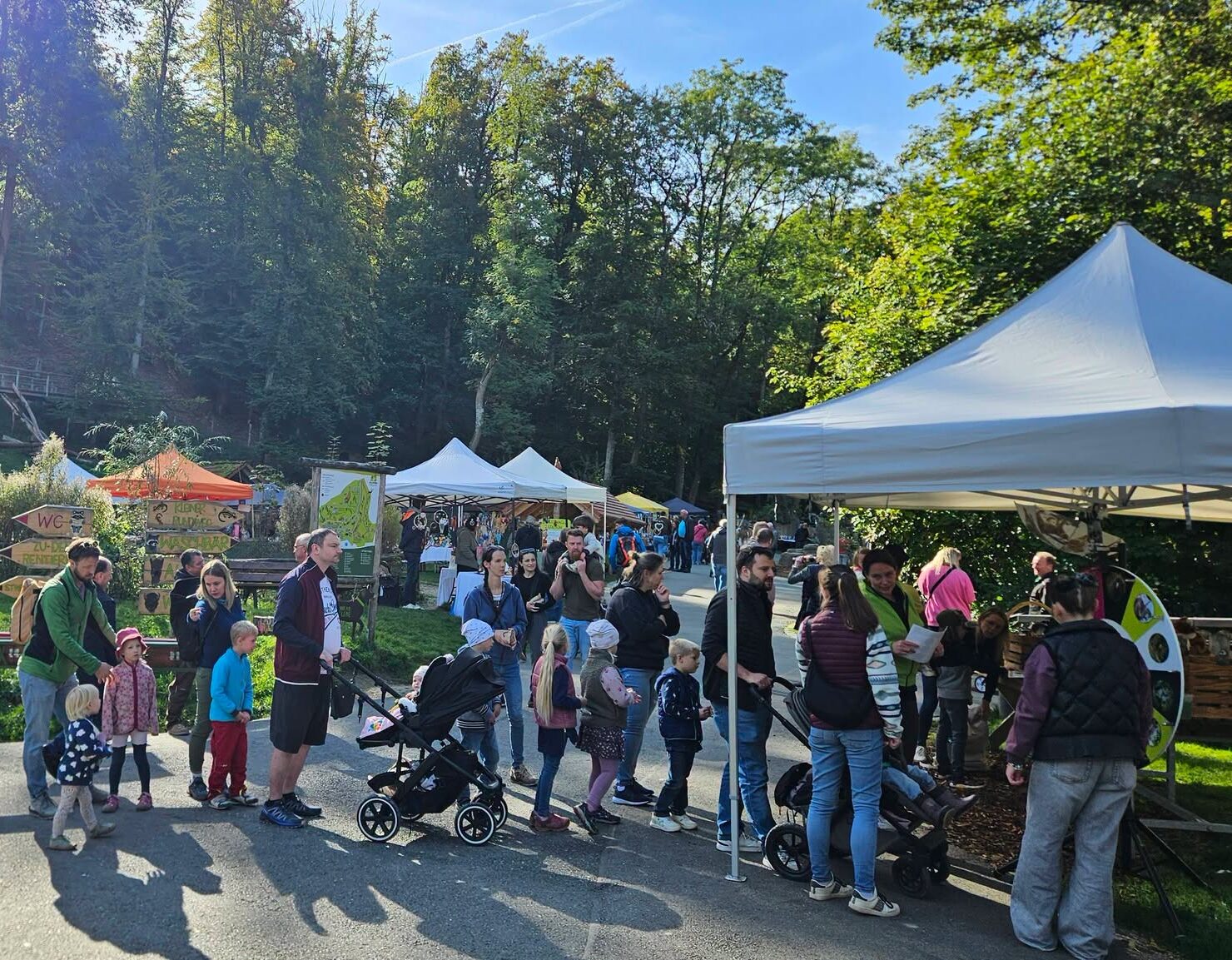 Viele Menschen versammeln sich bei sonnigem Wetter auf einer Wiese mit Ständen und Zelten, umgeben von Bäumen und Wald.